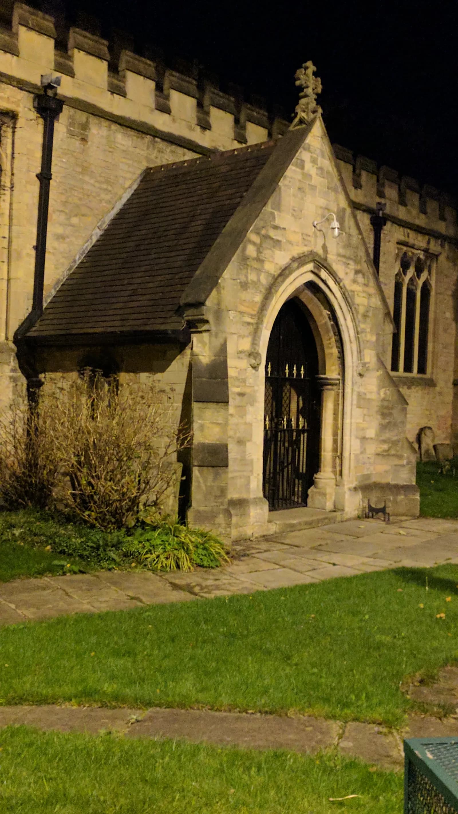 A stone church entrance with an arched doorway and iron gate, surrounded by green grass and subtle lighting at night.